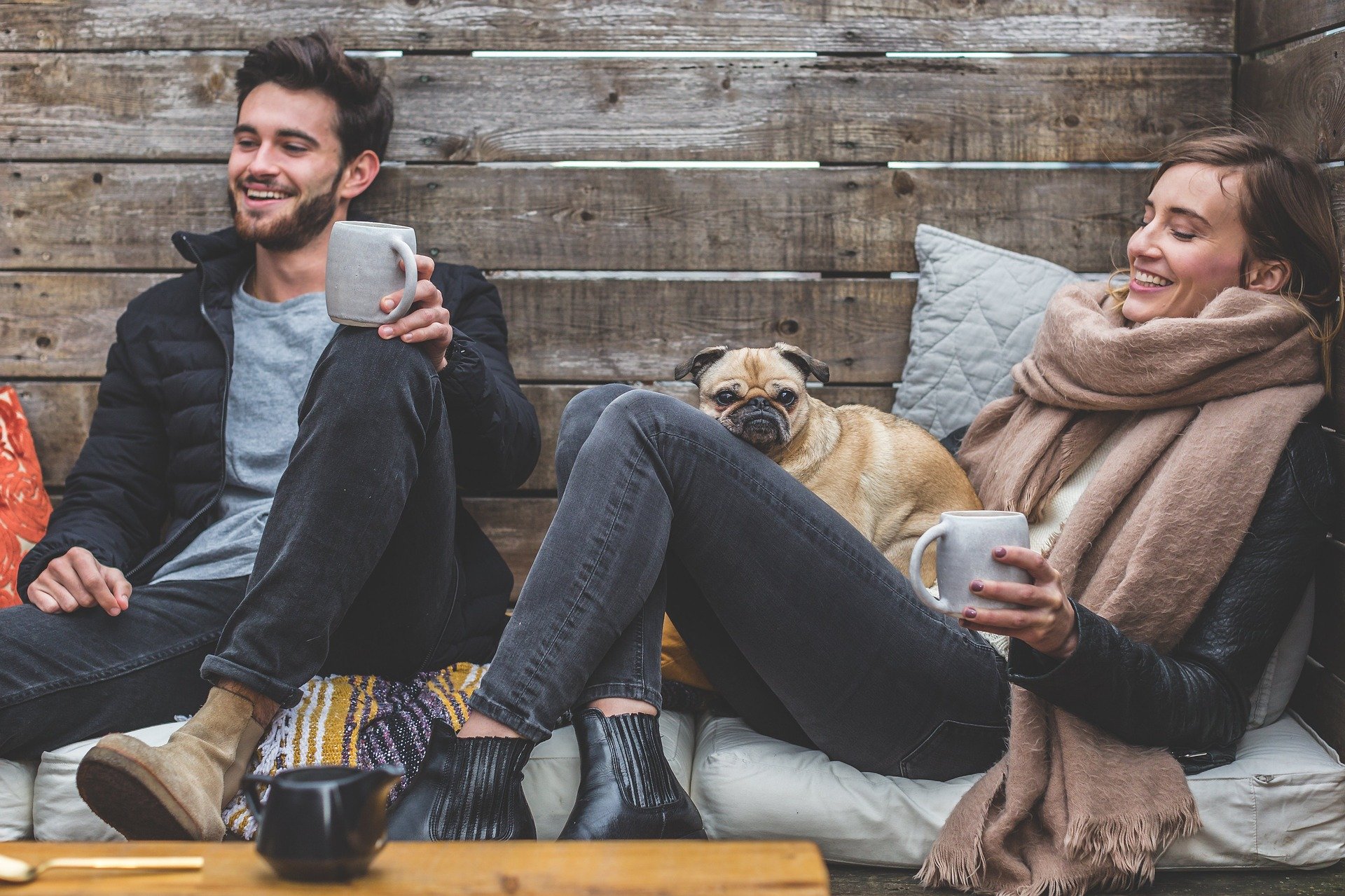 Zwei junge Menschen mit Kaffeebecher in den Händen und einem Hund an der Seite, die gemeinsam am Tisch abhängen