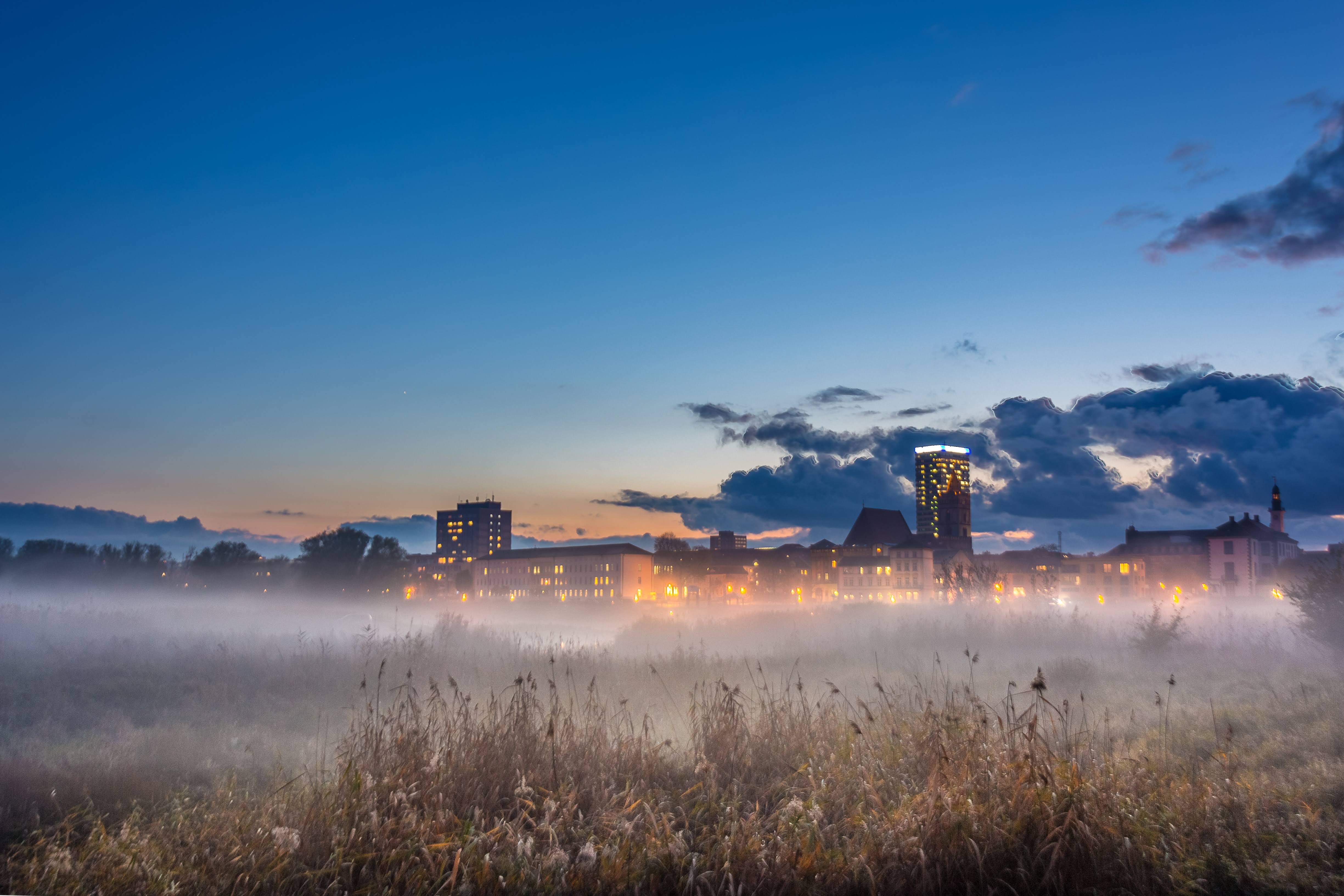 Panorama von der Stadt in der Nacht, im Vordergrund eine Wiese mit Nebel. Oderturm leuchtet im Hintergrund.