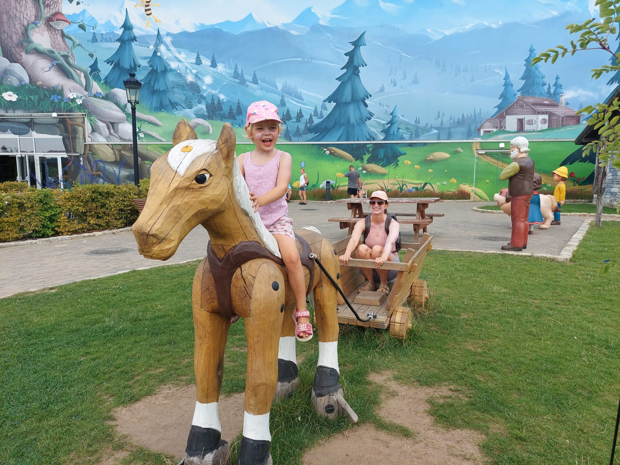EIn Mädchen sitzt auf dem Holzpferd im Majaland Freizeitpark, eine Mutti sitzt im Anhänger aus Holz. Beide freuen sich