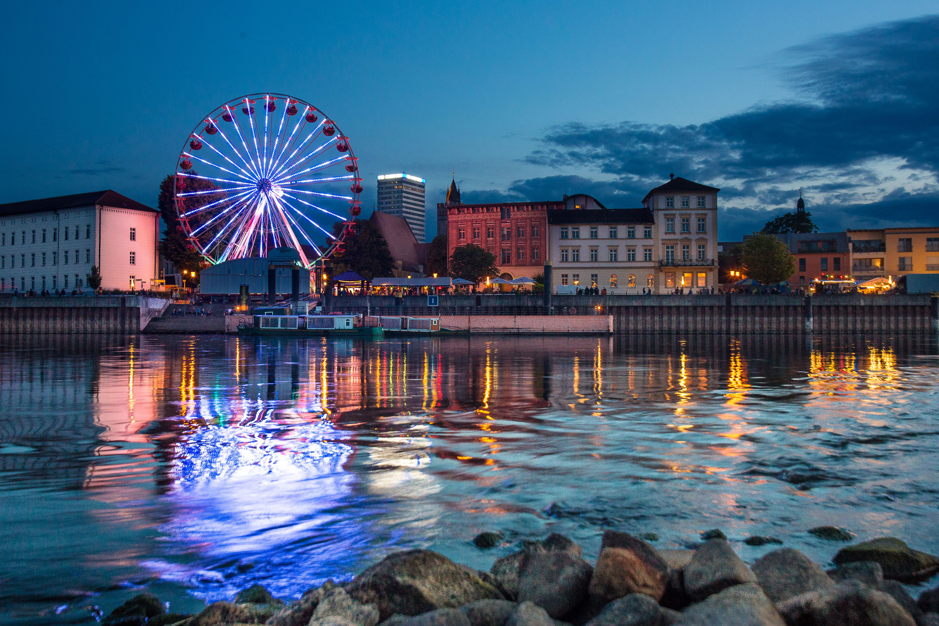 Nachtpanorama vom Uferpromenade der Stadt Frankfurt (Oder). Ein Riesenrad ist violett beleuchtet. Oderturm Schrift auf dem Oderturm leuchtet auch. Im Vordergrund einige Steine im Wasser