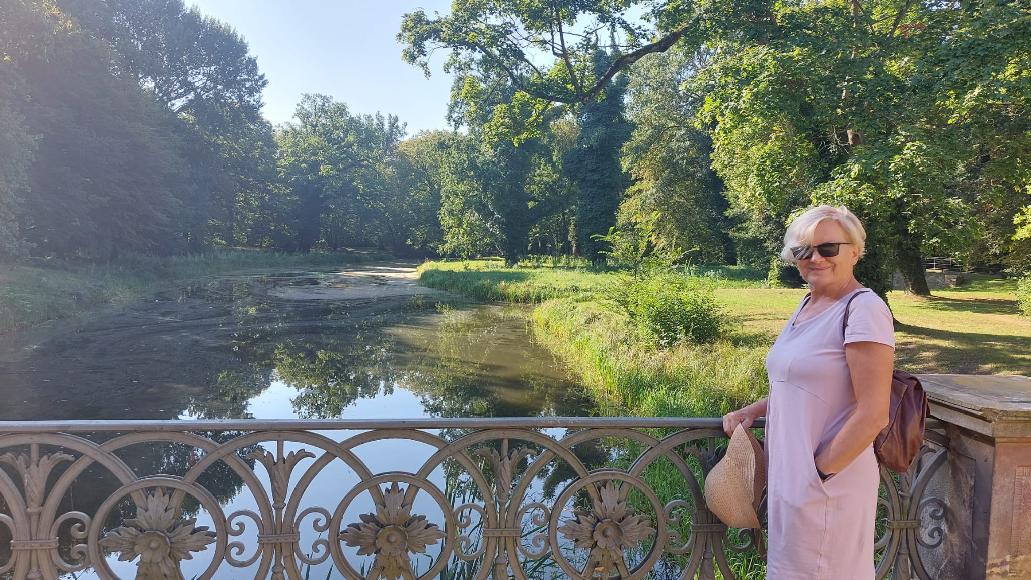 Eine Frau im Sommerkleid und Hut steht an der Brücke im Schlossgarten und lächelt.