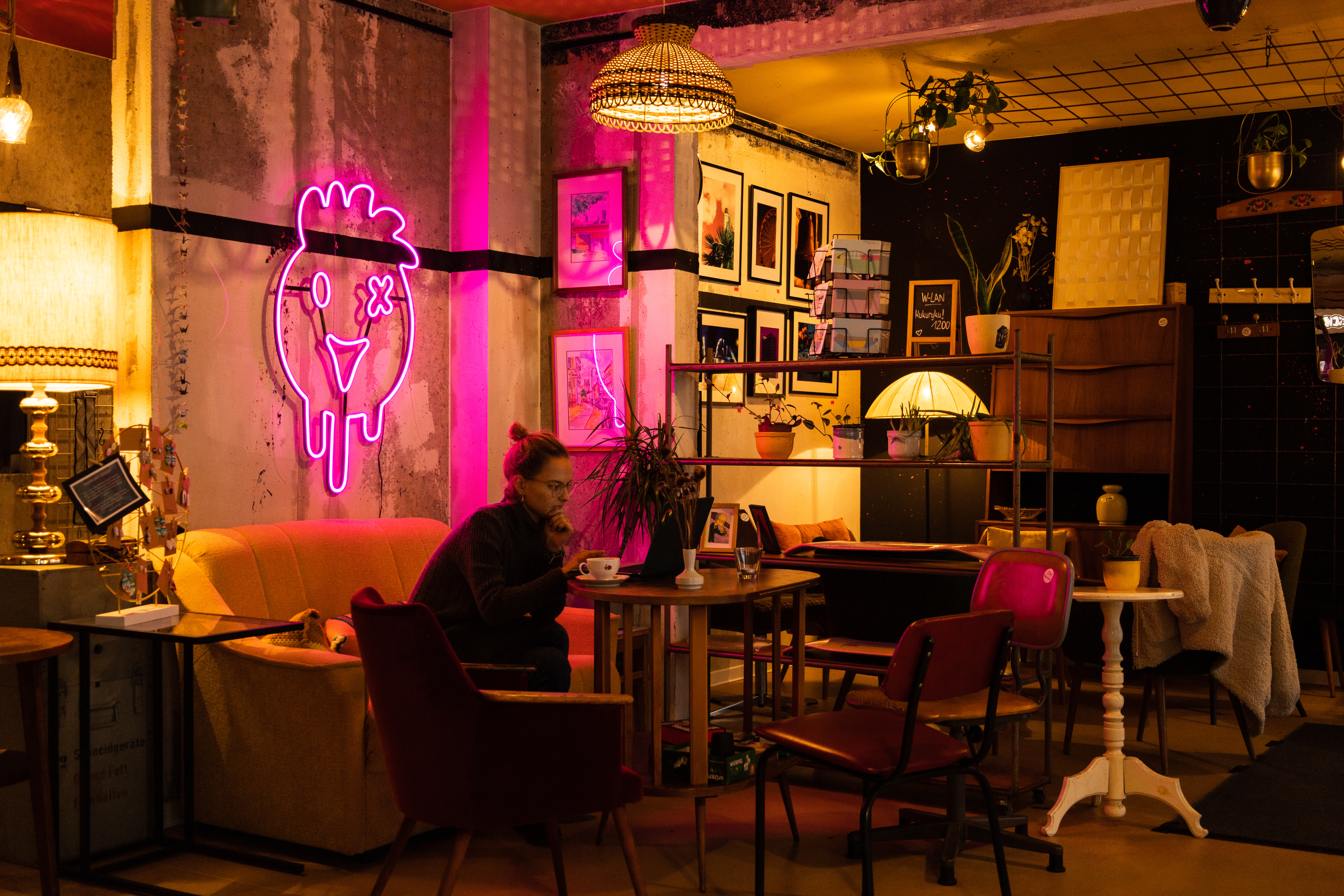 Eine Frau sitzt im stimmungsvollem Zimmer im Cafe, trinkt Cafe und arbeitet am Laptop. Sie sitzt auf dem Sofa. An der Wand hängt eine rosane Lampe im Form von Hahn.
